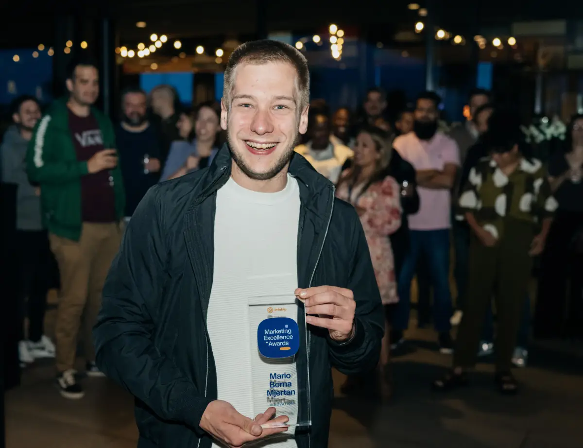 A picture of Mario Borna in 2024 on an event, holding an award plaque from Infobip Marketing Excellence Awards in the Innovation category. He is smiling. Behind him, a number of people stand, blurred.