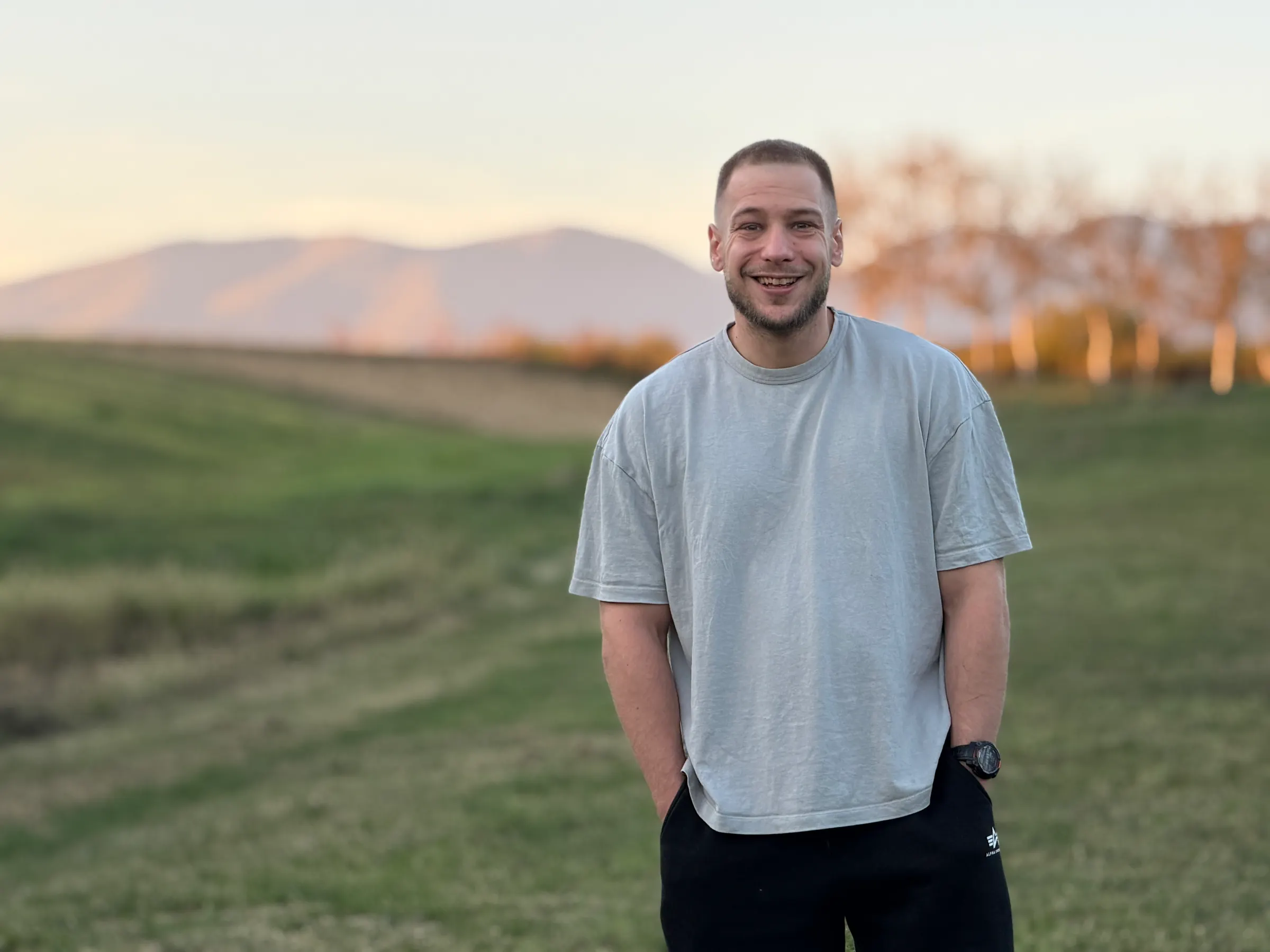 Borna standing in a grassy field at sunset, hands in pockets, hills behind him.