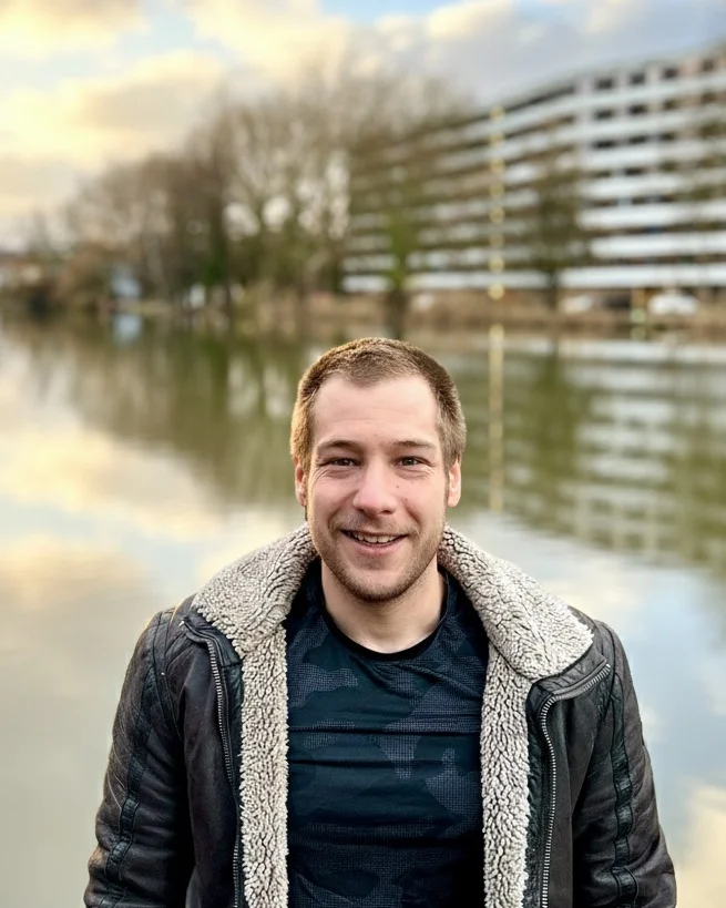 Borna smiling in front of a river at dusk, wearing a leather jacket with a sheepskin collar.